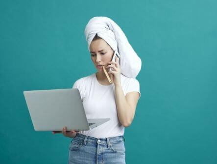 A woman with a towel around her hair is looking at her laptop with a toothbrush in her mouth